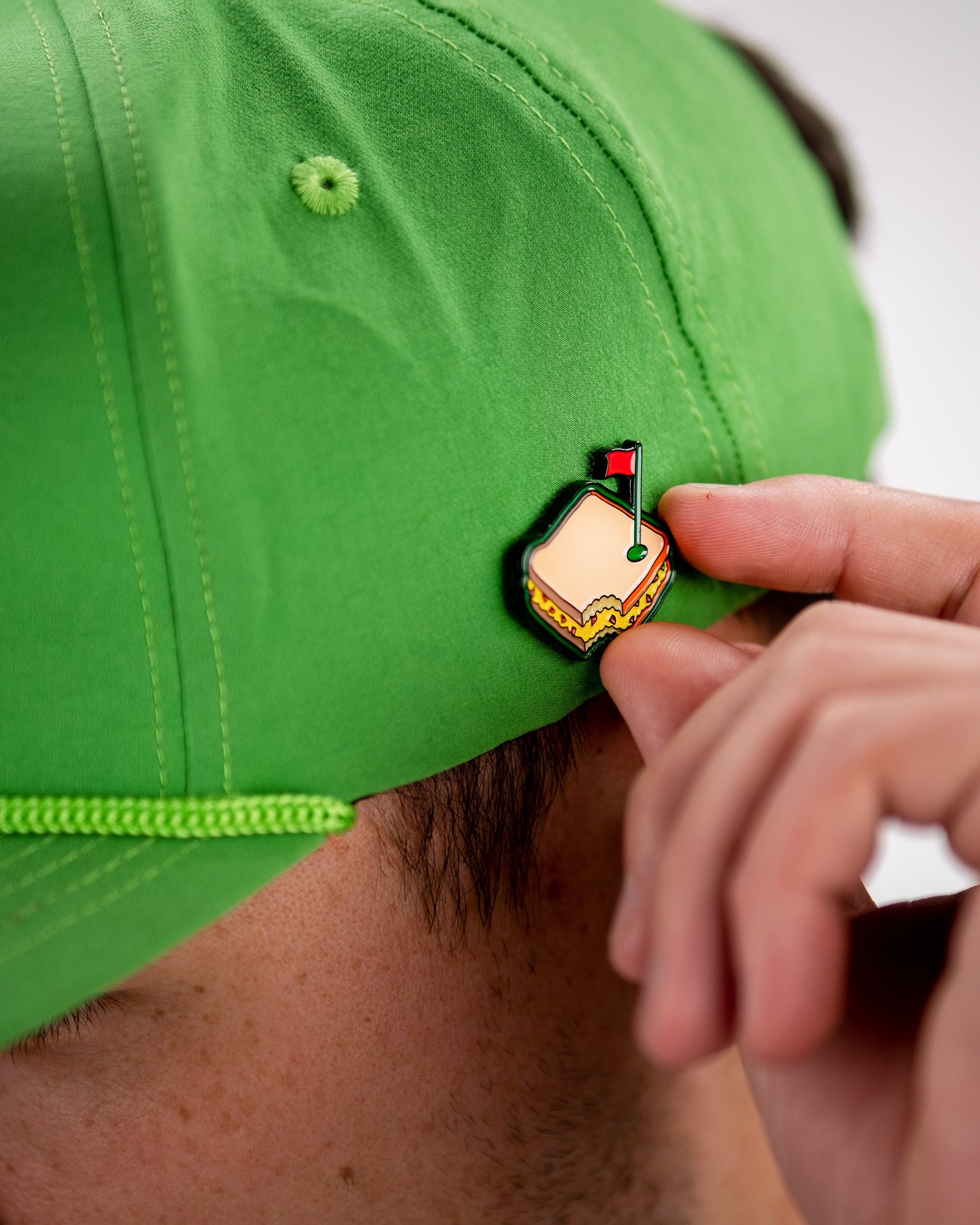 Someone wearing a bright green cap adjusts a Pins and Aces Pimento Cheese Sandwich Ball Marker enamel pin, shaped like an Augusta golf marker with a red flag, attached to the back of the cap.