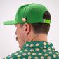 A person wearing a green cap with a Pins and Aces Pimento Cheese Sandwich Ball Marker attached, along with a patterned green shirt featuring sandwiches, shown from behind with their face partially visible.