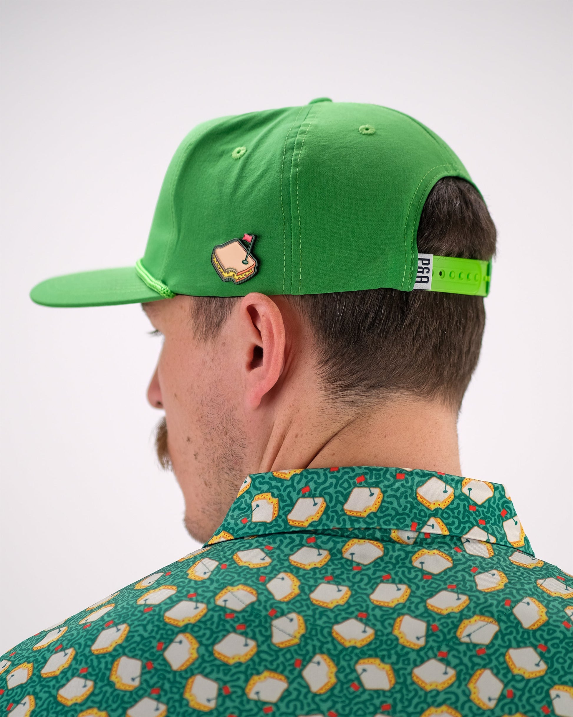 A person wearing a green cap with a Pins and Aces Pimento Cheese Sandwich Ball Marker attached, along with a patterned green shirt featuring sandwiches, shown from behind with their face partially visible.