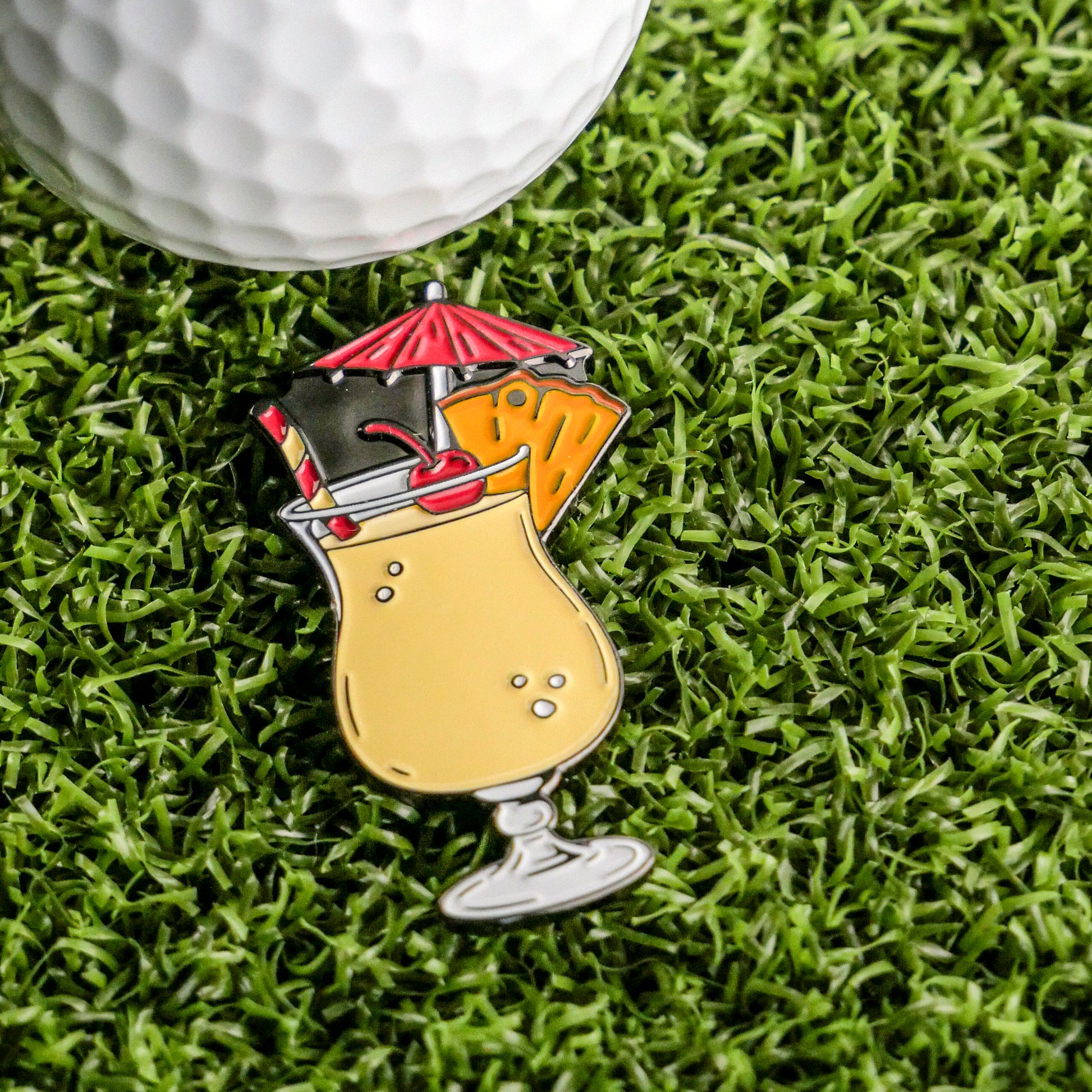 The Piña Colada Ball Marker by Ballmarkers.com, a vibrant enamel pin featuring a straw, umbrella, and fruit garnish, is shown on green grass next to a white golf ball.