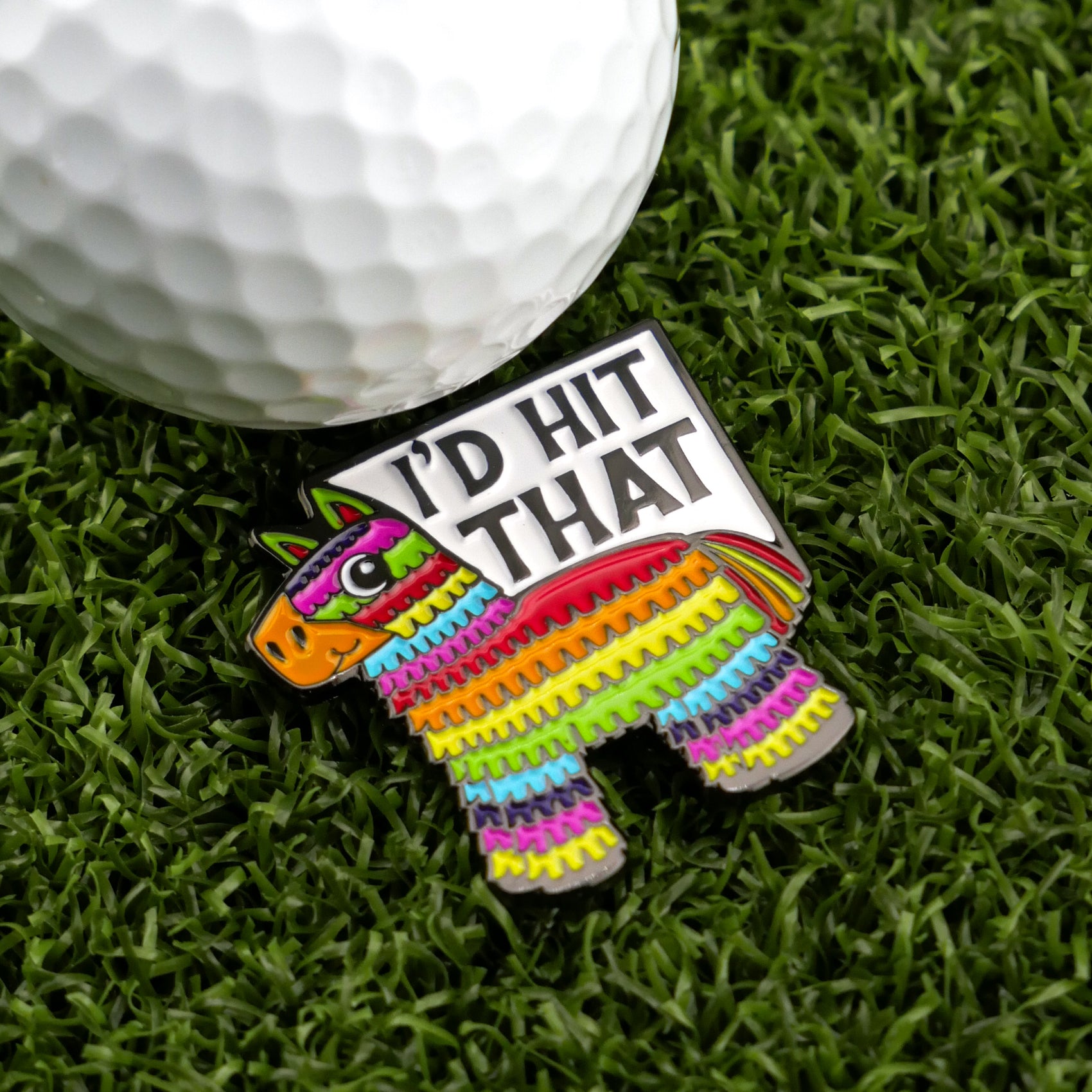 The Logie Joe's Golf "I’d Hit That Piñata" pin is a colorful, piñata-shaped golf ball marker with funny text, shown on artificial grass next to a white golf ball—perfect for anyone who enjoys humorous golf accessories.