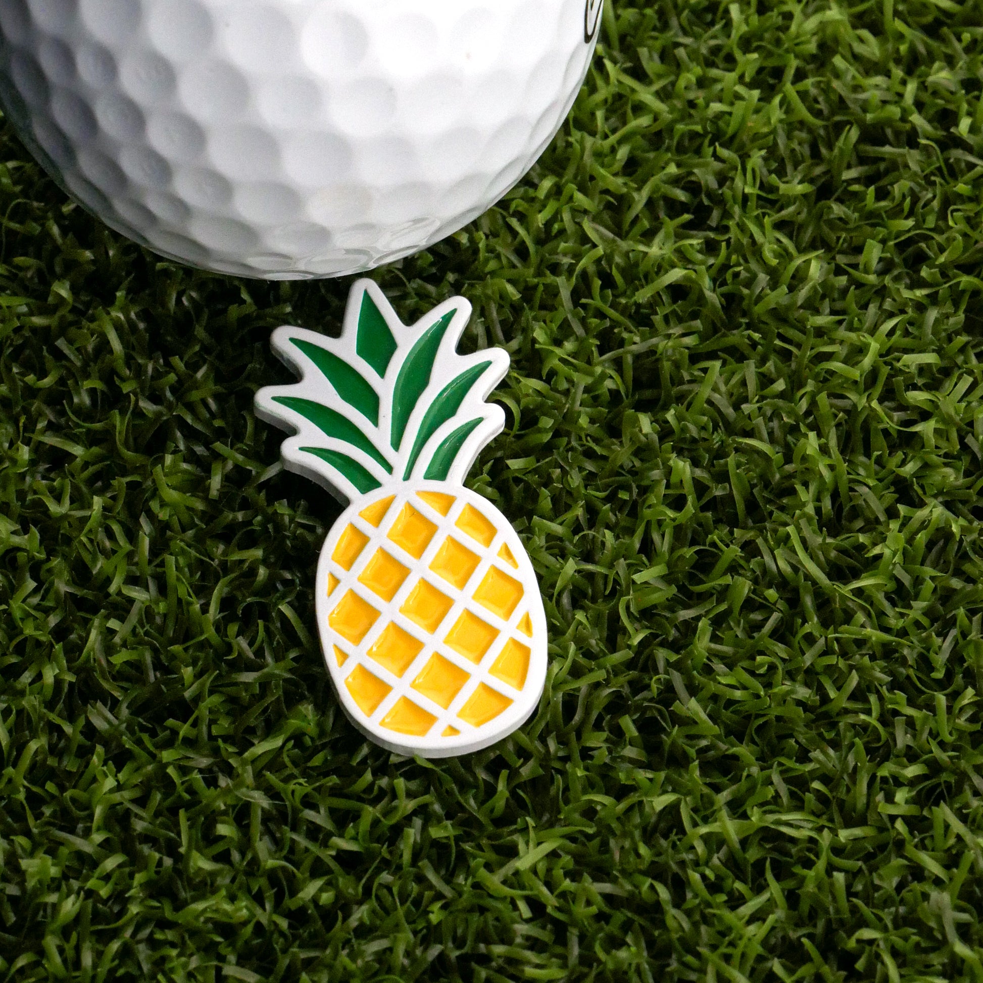 A Logie Joe’s Golf Pineapple Ball Marker, featuring green leaves and a yellow crisscross pattern, adds flair alongside a white golf ball as they rest on green artificial grass.
