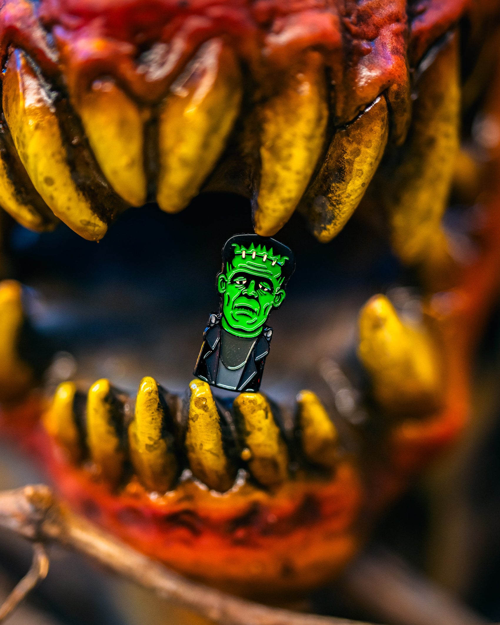 A Pins and Aces Frankenstein Ball Marker is gripped between the sharp yellow teeth of a fierce, red-mouthed creature with large fangs, creating a dramatic scene emphasized by the blurred background—an eye-catching golf accessory in action.