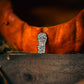 The Pins and Aces Mummy Ball Marker, a small enamel pin shaped like a mummy, is displayed before a carved pumpkin on a wooden surface with a dark and moody background.