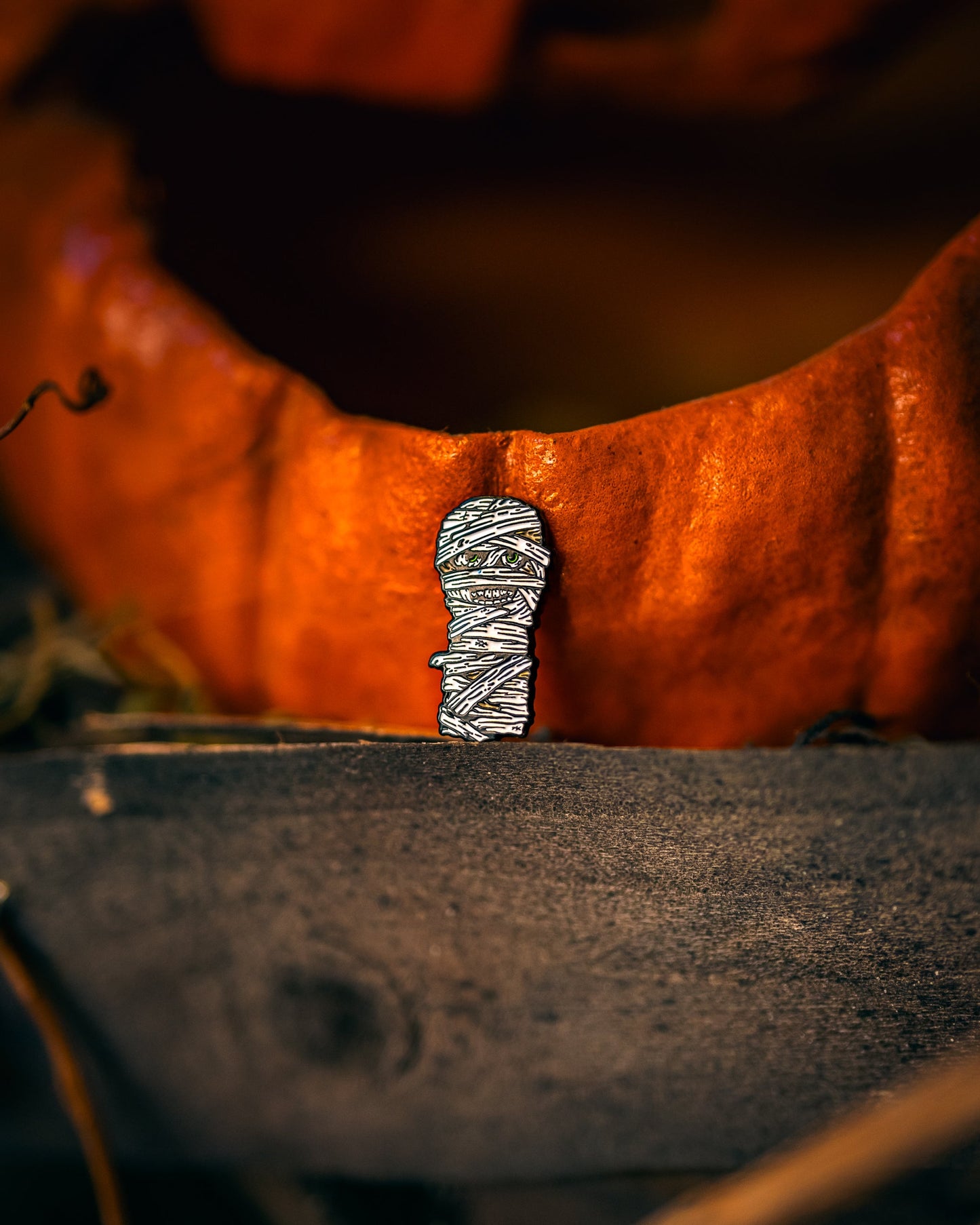 The Pins and Aces Mummy Ball Marker, a small enamel pin shaped like a mummy, is displayed before a carved pumpkin on a wooden surface with a dark and moody background.