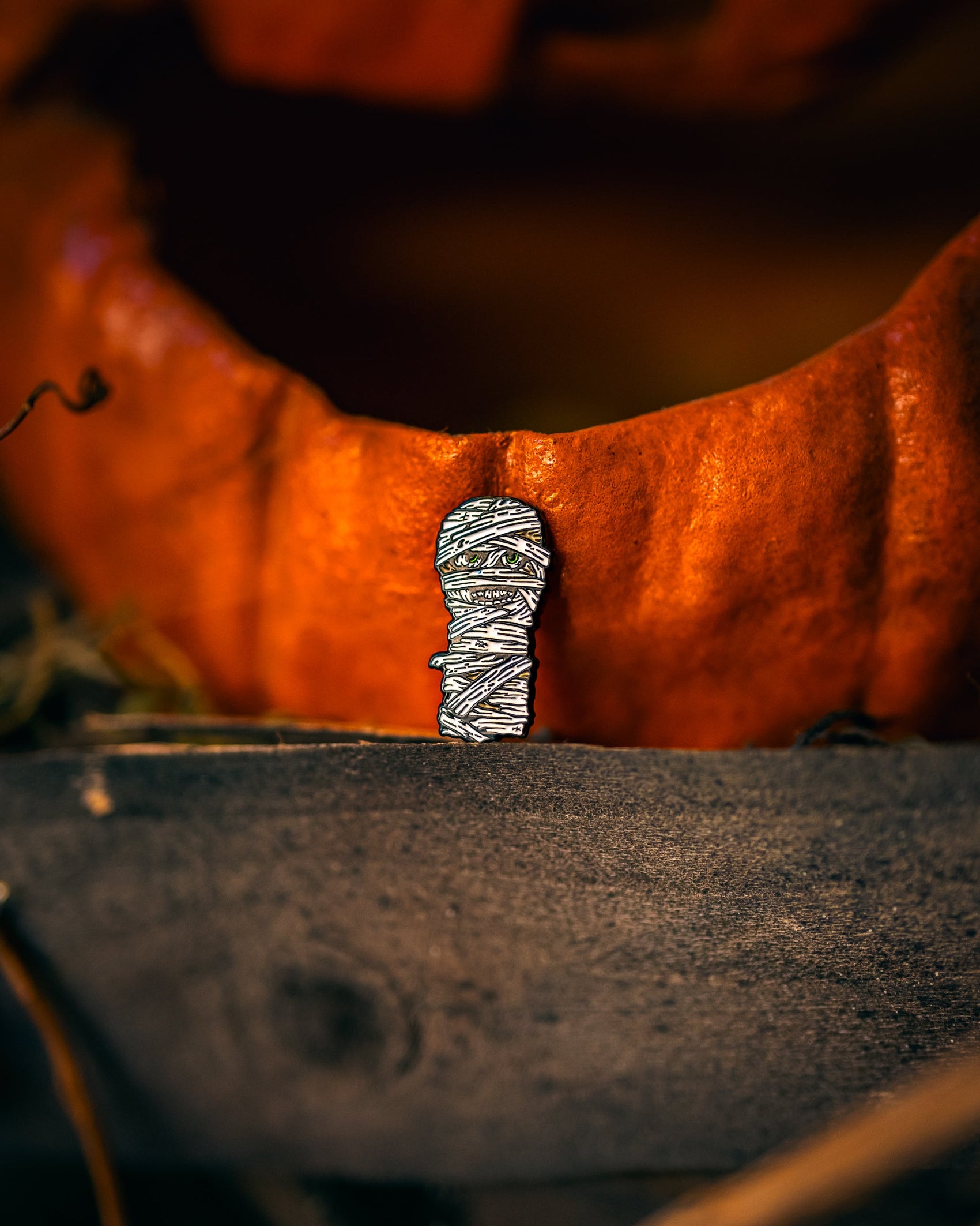 The Pins and Aces Mummy Ball Marker, a small enamel pin shaped like a mummy, is displayed before a carved pumpkin on a wooden surface with a dark and moody background.