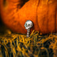 The Skeleton Ball Marker by Pins and Aces, shaped like a skull with a spine, is set on a textured yellow surface. Blurred orange pumpkins in the background create a festive Halloween or autumn vibe.