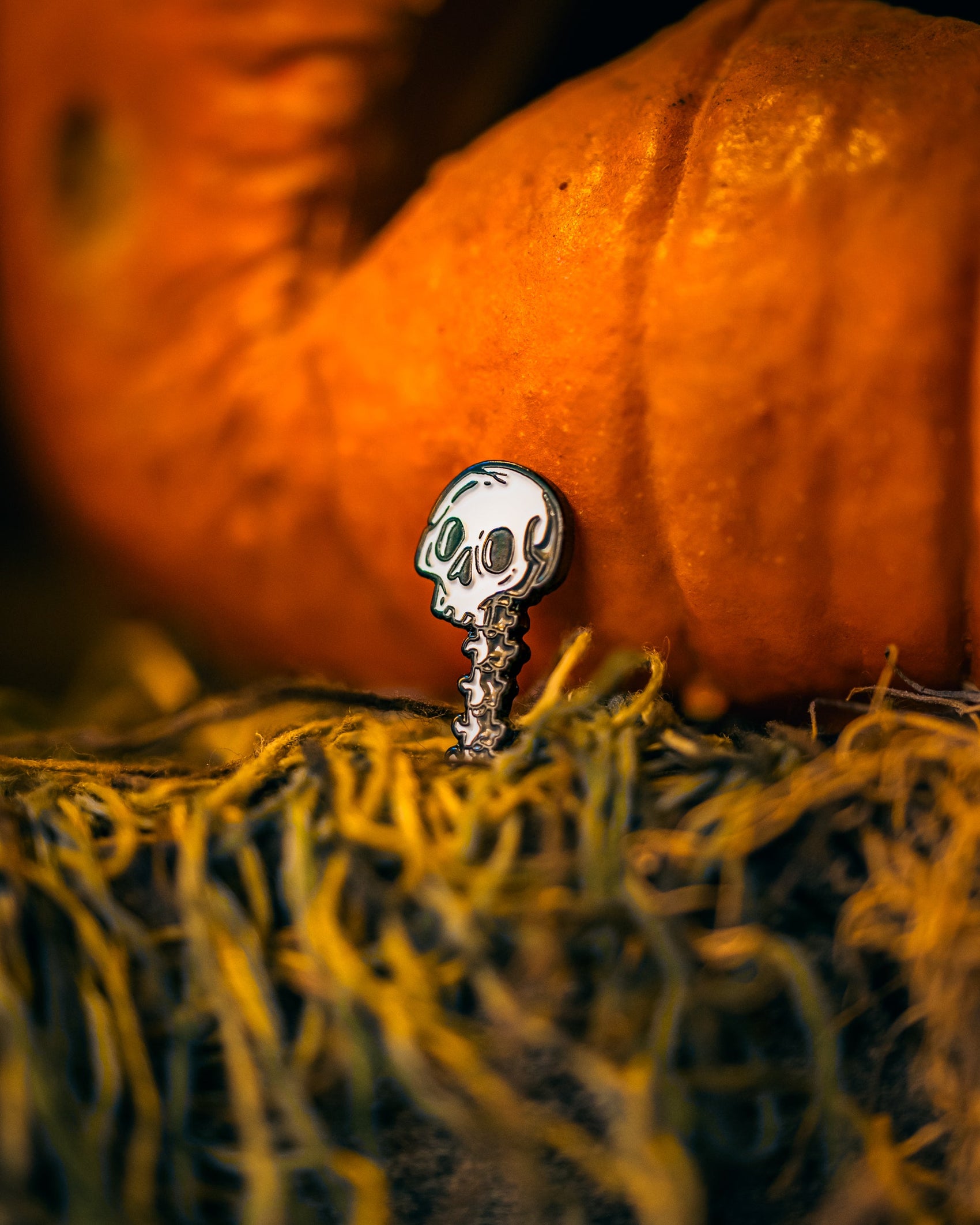 The Skeleton Ball Marker by Pins and Aces, shaped like a skull with a spine, is set on a textured yellow surface. Blurred orange pumpkins in the background create a festive Halloween or autumn vibe.