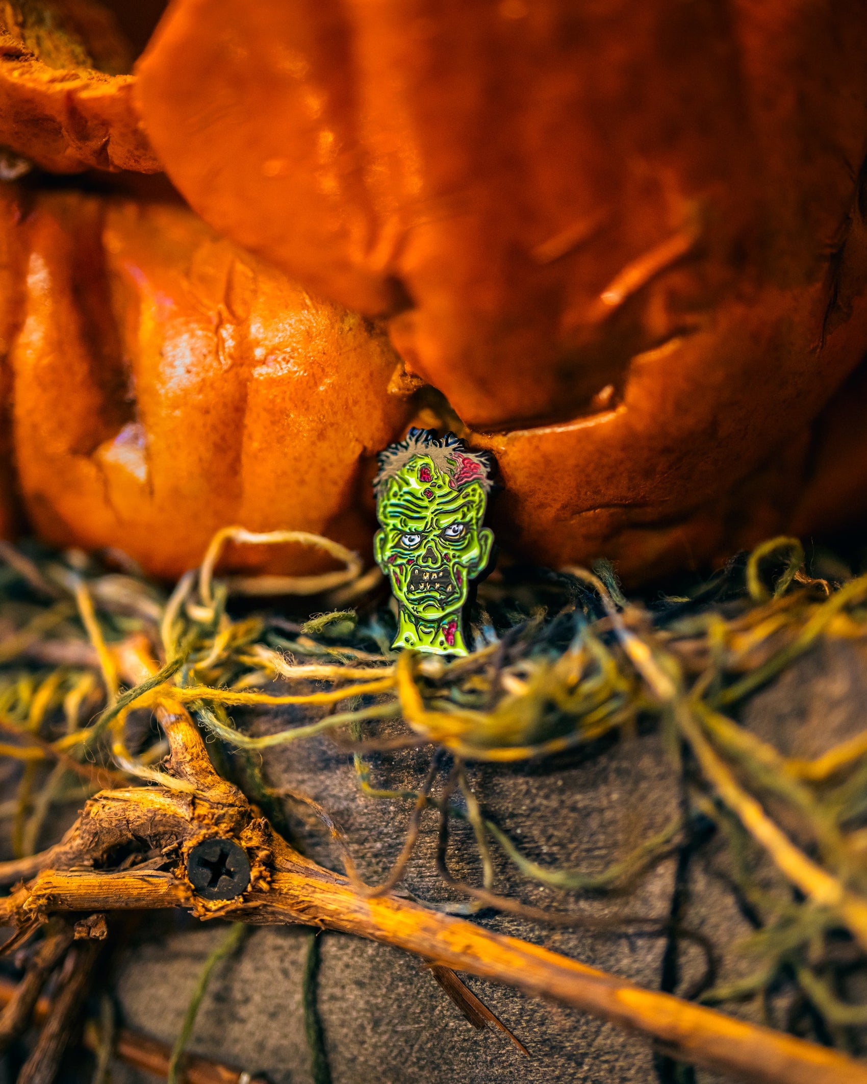 The Pins and Aces Mutant Zombie - Ball Marker, in bright green, sits among yarn and twigs beneath a large orange jack-o’-lantern’s mouth, creating a spooky Halloween vibe.