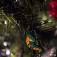 A close-up of the Pins and Aces T-Rex Ornament, a quirky green and orange dinosaur Christmas golf accessory, hanging from a tree branch among dark green needles and festive lights.
