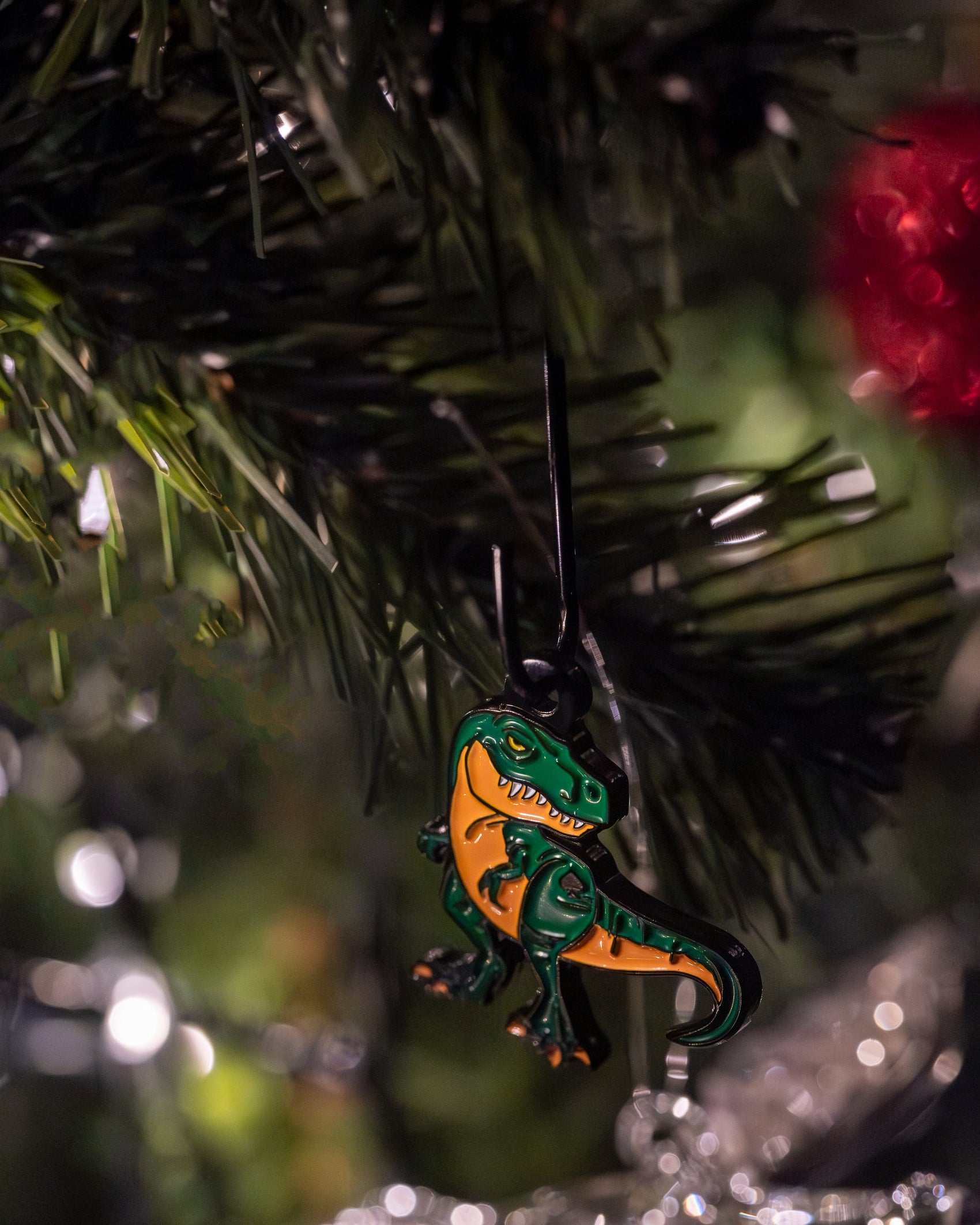 A close-up of the Pins and Aces T-Rex Ornament, a quirky green and orange dinosaur Christmas golf accessory, hanging from a tree branch among dark green needles and festive lights.