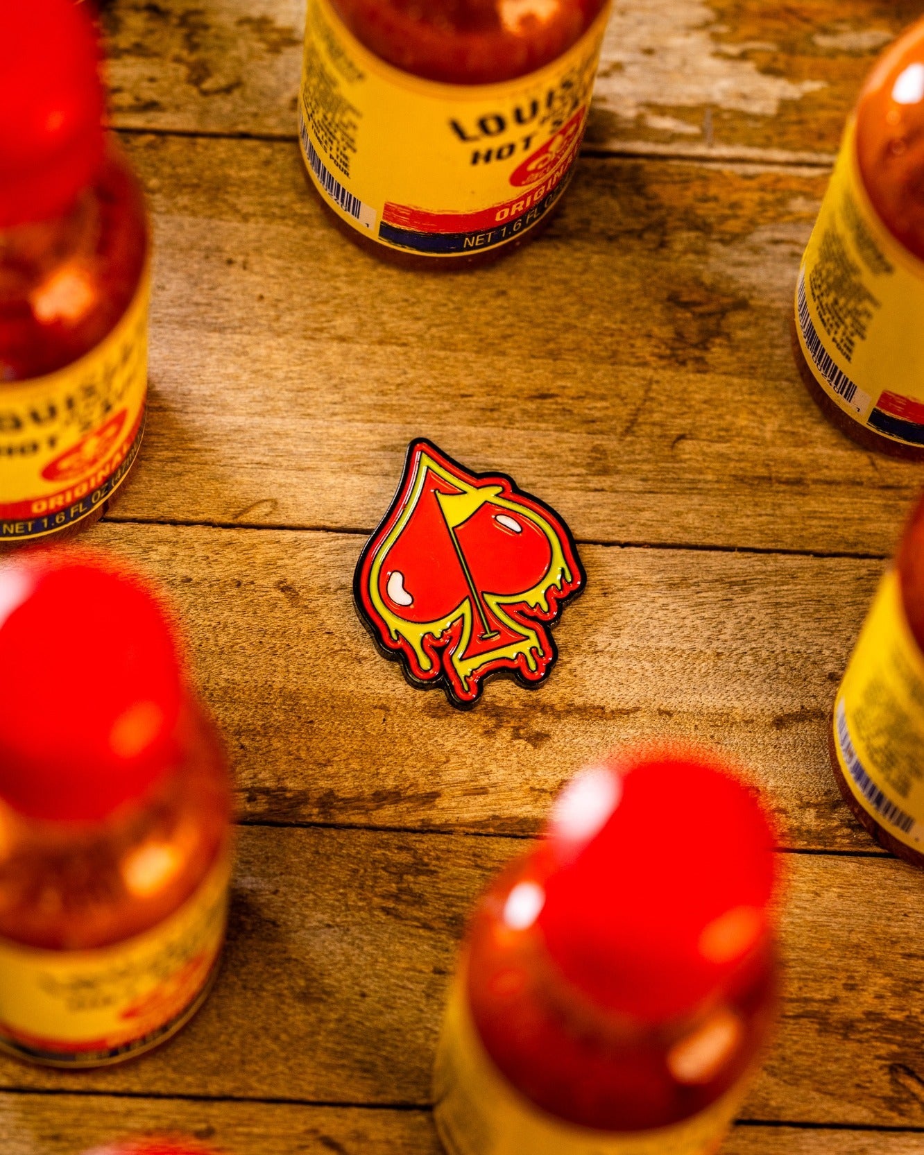 A Pins and Aces Flamin' Spade Ball Marker sits on a wooden surface near several Louisiana hot sauce bottles with red caps, alongside a patch featuring a dripping red spade design.