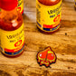 Three bottles of Louisiana hot sauce rest on a rustic wooden surface next to the Flamin' Spade Ball Marker by Pins and Aces, a red and yellow patch shaped like a spade with dripping details.