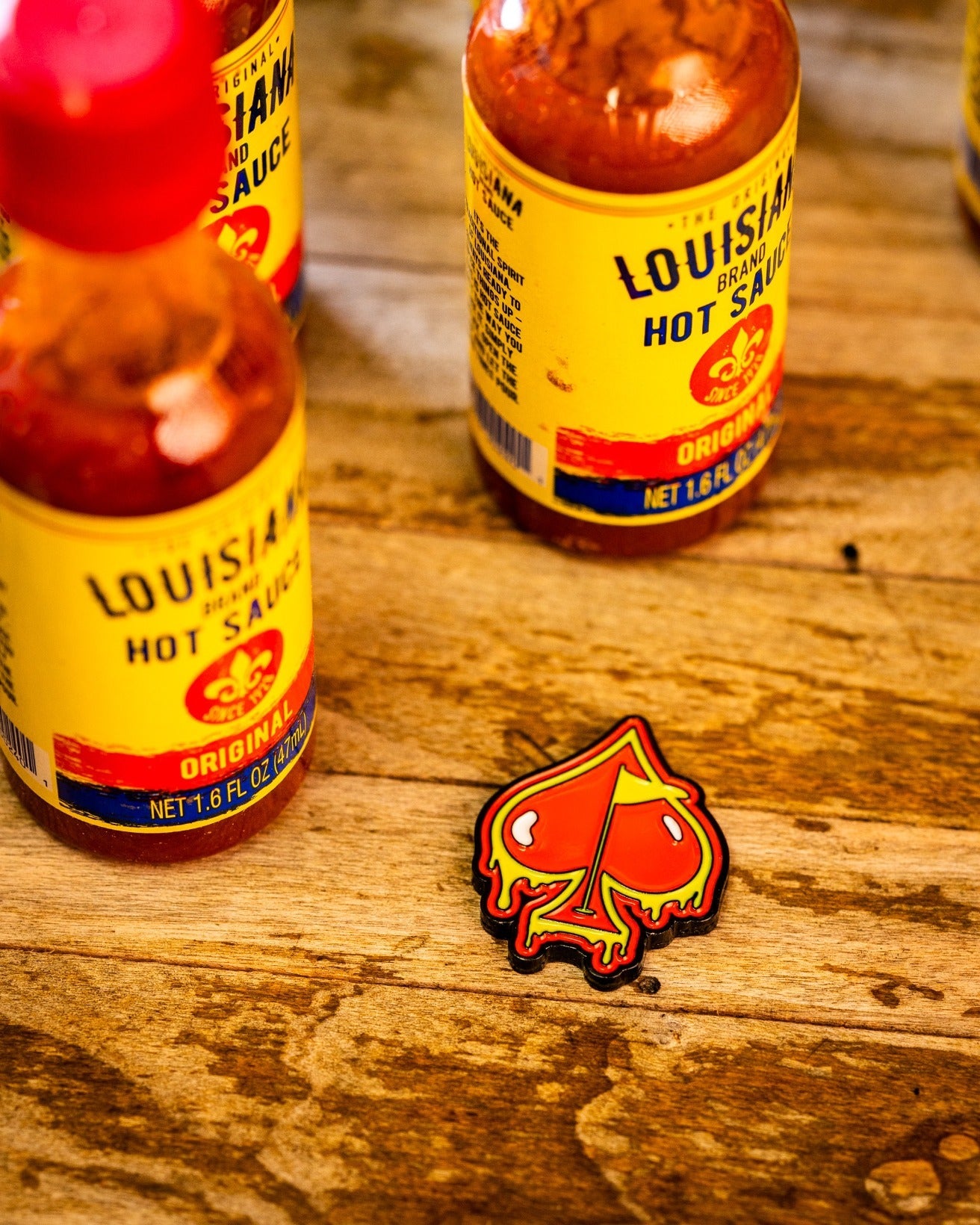 Three bottles of Louisiana hot sauce rest on a rustic wooden surface next to the Flamin' Spade Ball Marker by Pins and Aces, a red and yellow patch shaped like a spade with dripping details.