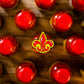 Rows of bottles with bright red caps seen from above, one in the center topped with Pins and Aces’ Fleur-de-lis Ball Marker—a premium golf accessory featuring a decorative yellow and red design. The background is wooden.