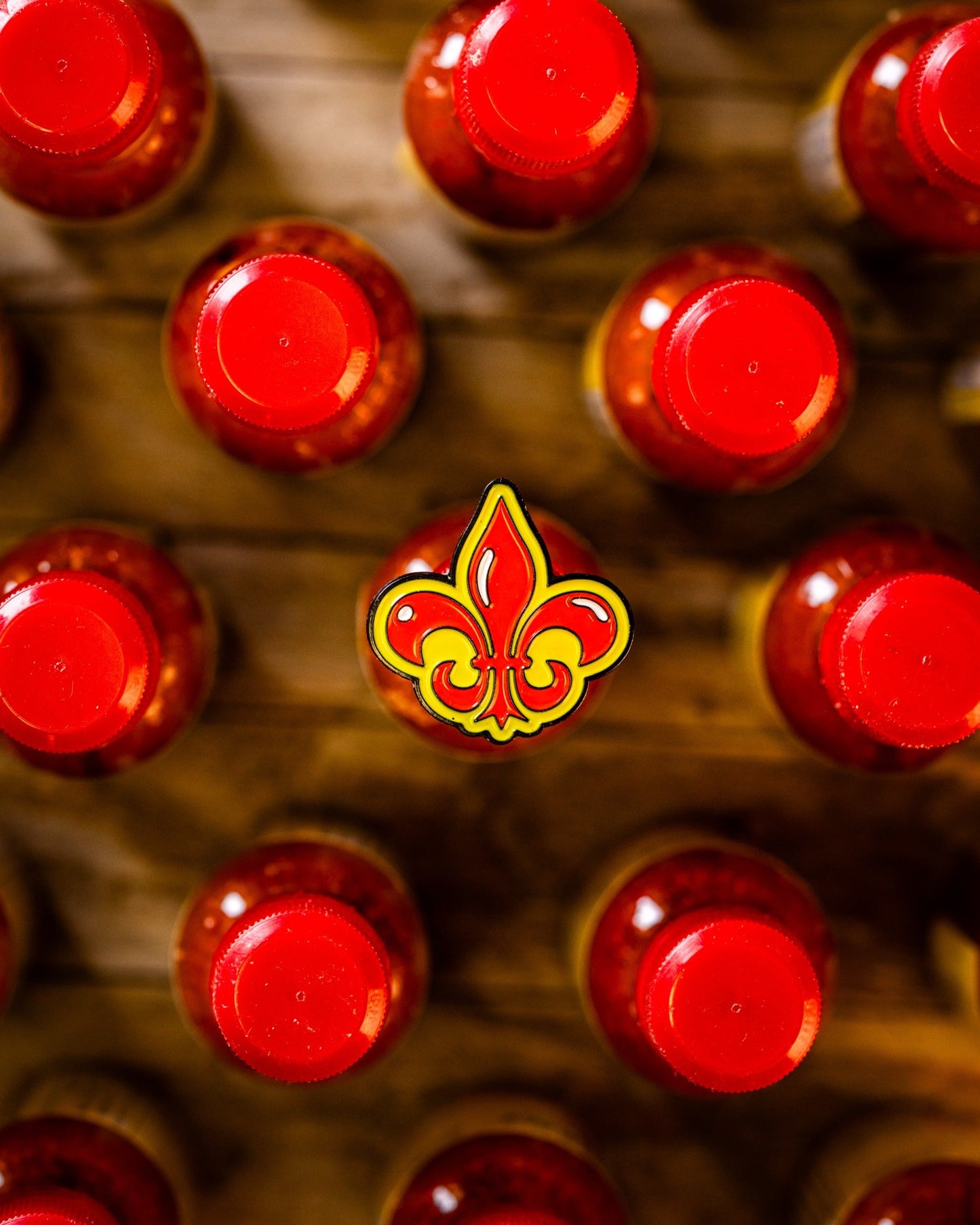 Rows of bottles with bright red caps seen from above, one in the center topped with Pins and Aces’ Fleur-de-lis Ball Marker—a premium golf accessory featuring a decorative yellow and red design. The background is wooden.