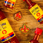 Four Louisiana hot sauce bottles with yellow labels sit on wood, framing two Pins and Aces enamel pins—a spade and the Fleur-de-lis Ball Marker—ideal as premium golf accessories or magnetic hat clips.
