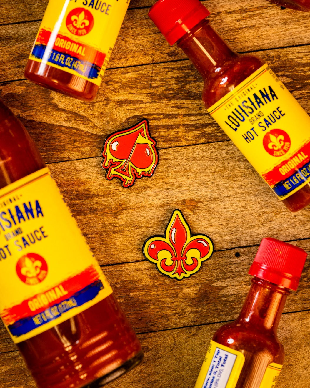 Four Louisiana hot sauce bottles with yellow labels sit on wood, framing two Pins and Aces enamel pins—a spade and the Fleur-de-lis Ball Marker—ideal as premium golf accessories or magnetic hat clips.