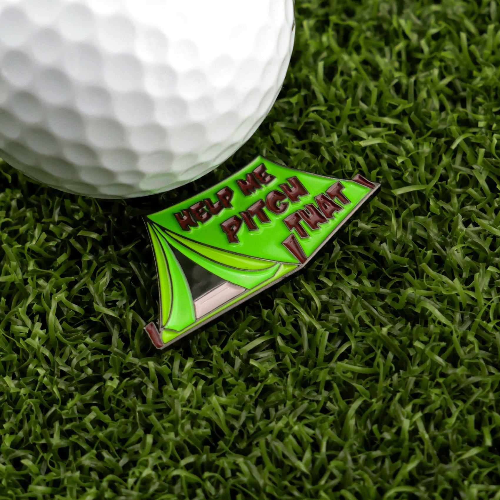 A close-up of a golf ball on green grass beside Logie Joe's Golf "Pitch That Tent" marker, shaped like a tent and inscribed with HELP ME PITCH THAT TENT.