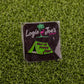 The Pitch That Tent ball marker from Logie Joe's Golf features a green tent and "HELP ME PITCH THAT" on artificial grass. Behind it, a black card with a pink flamingo, palm tree, and Logie Joe’s Golf adds flair to this fun accessory.