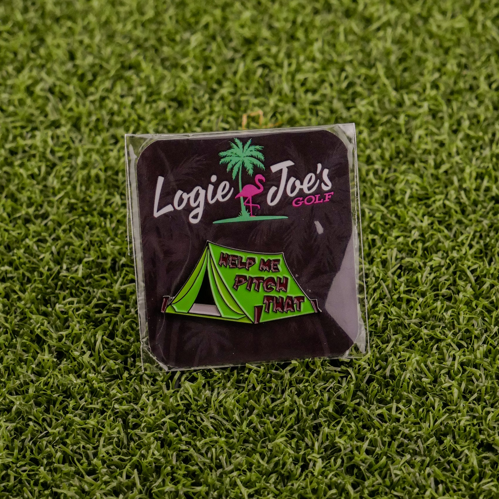 The Pitch That Tent ball marker from Logie Joe's Golf features a green tent and "HELP ME PITCH THAT" on artificial grass. Behind it, a black card with a pink flamingo, palm tree, and Logie Joe’s Golf adds flair to this fun accessory.