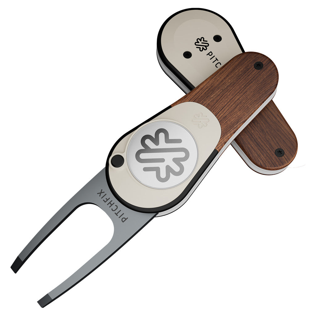 The PitchFix Woodie Divot Tool is an elegant metal fork with wood-effect styling, featuring the PitchFix logo and abstract symbol, shown with its cover partially removed.