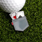 A golf ball on artificial grass sits next to the Ballmarkers.com Pocket Aces Ball Marker, a shiny grey poker-themed accessory holding the ace of hearts and ace of spades.