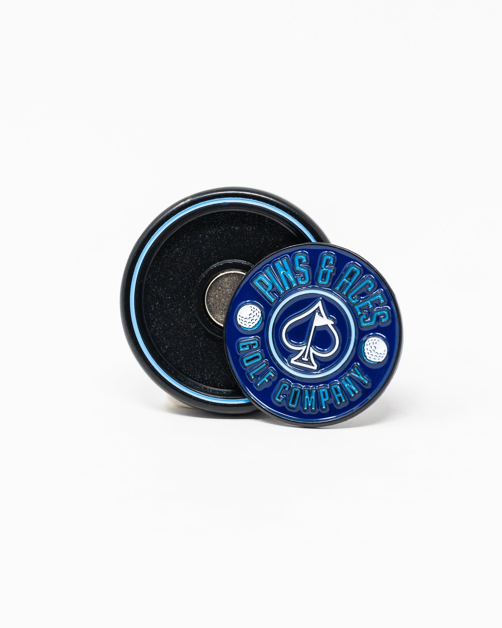 The Pop Out Medallion Ball Marker - Navy by Pins and Aces is a round, thin marker in blue and black with the brand name and ace of spades logo. It stands upright, showcasing its magnetic back on a white background.