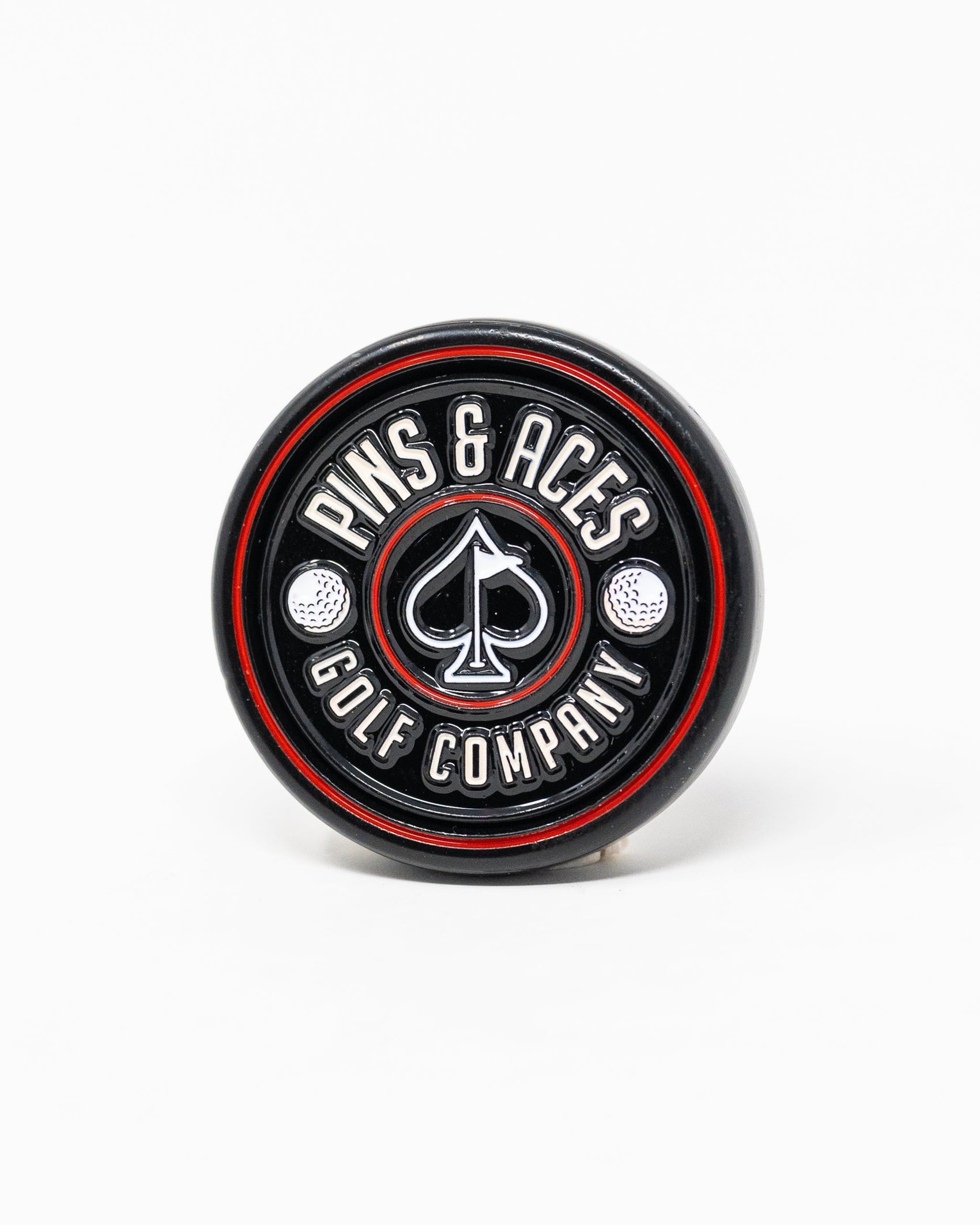 The Pop Out Medallion Ball Marker - Black by Pins and Aces is a thin, black circular marker with red and silver accents, featuring the brand name around a spade symbol and two golf balls.