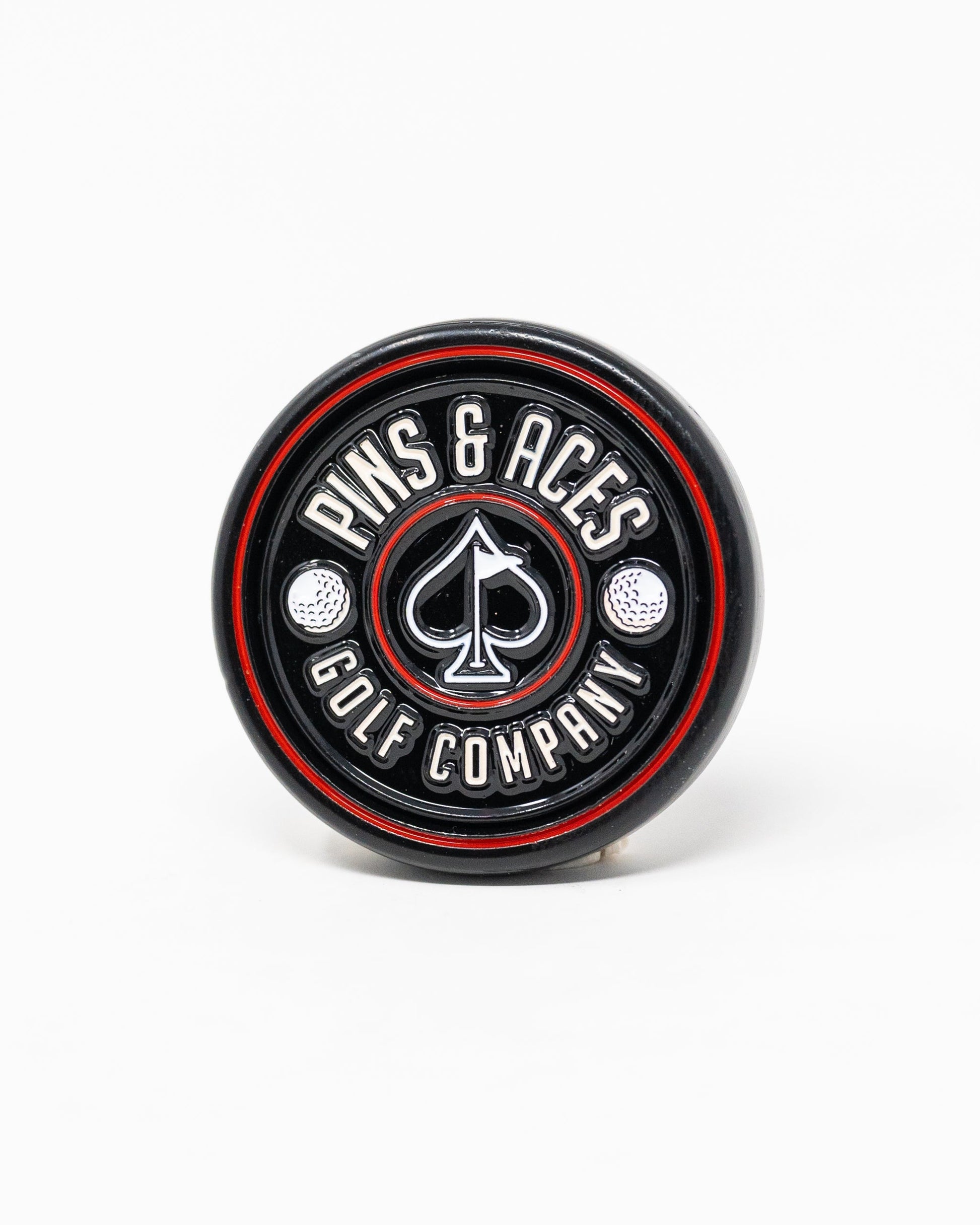 The Pop Out Medallion Ball Marker - Black by Pins and Aces is a thin, black circular marker with red and silver accents, featuring the brand name around a spade symbol and two golf balls.