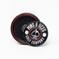 The Pop Out Medallion Ball Marker - Black by Pins and Aces features a round, black, magnetic marker with white branding, two golf balls, and an ace of spades symbol. It includes a thin marker displayed in front of its round holder with a red ring.