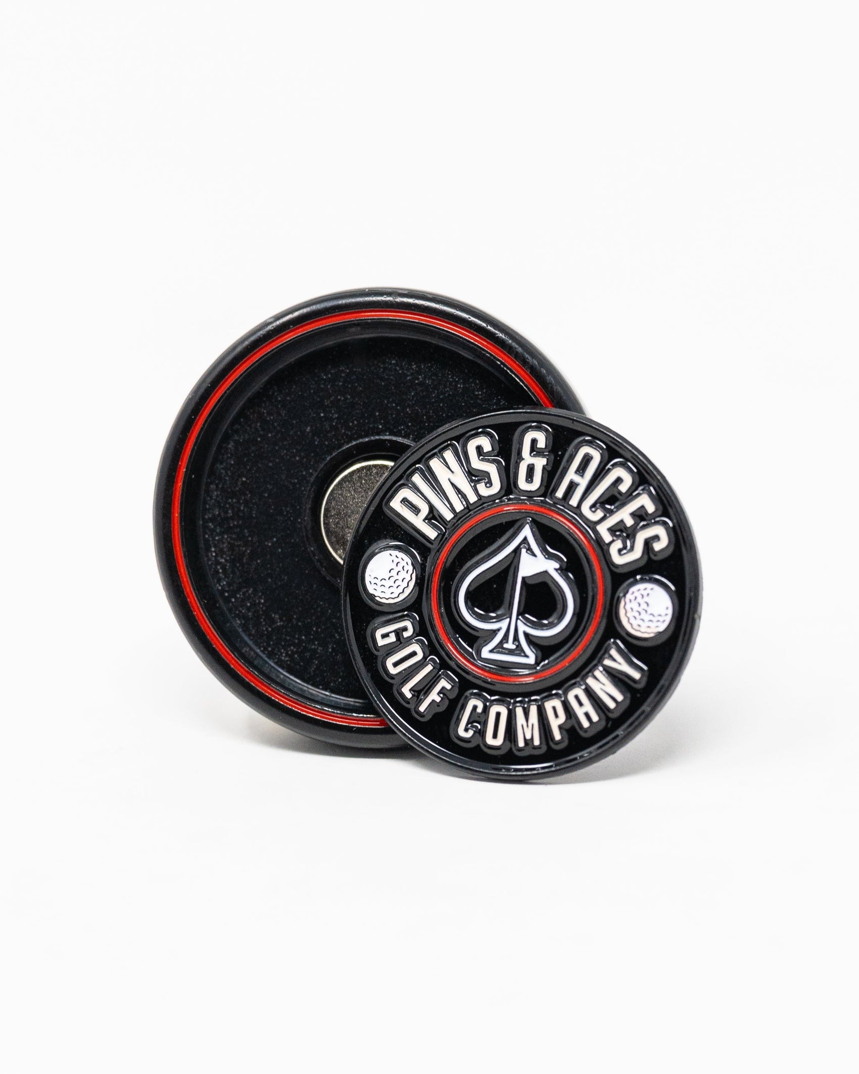 The Pop Out Medallion Ball Marker - Black by Pins and Aces features a round, black, magnetic marker with white branding, two golf balls, and an ace of spades symbol. It includes a thin marker displayed in front of its round holder with a red ring.