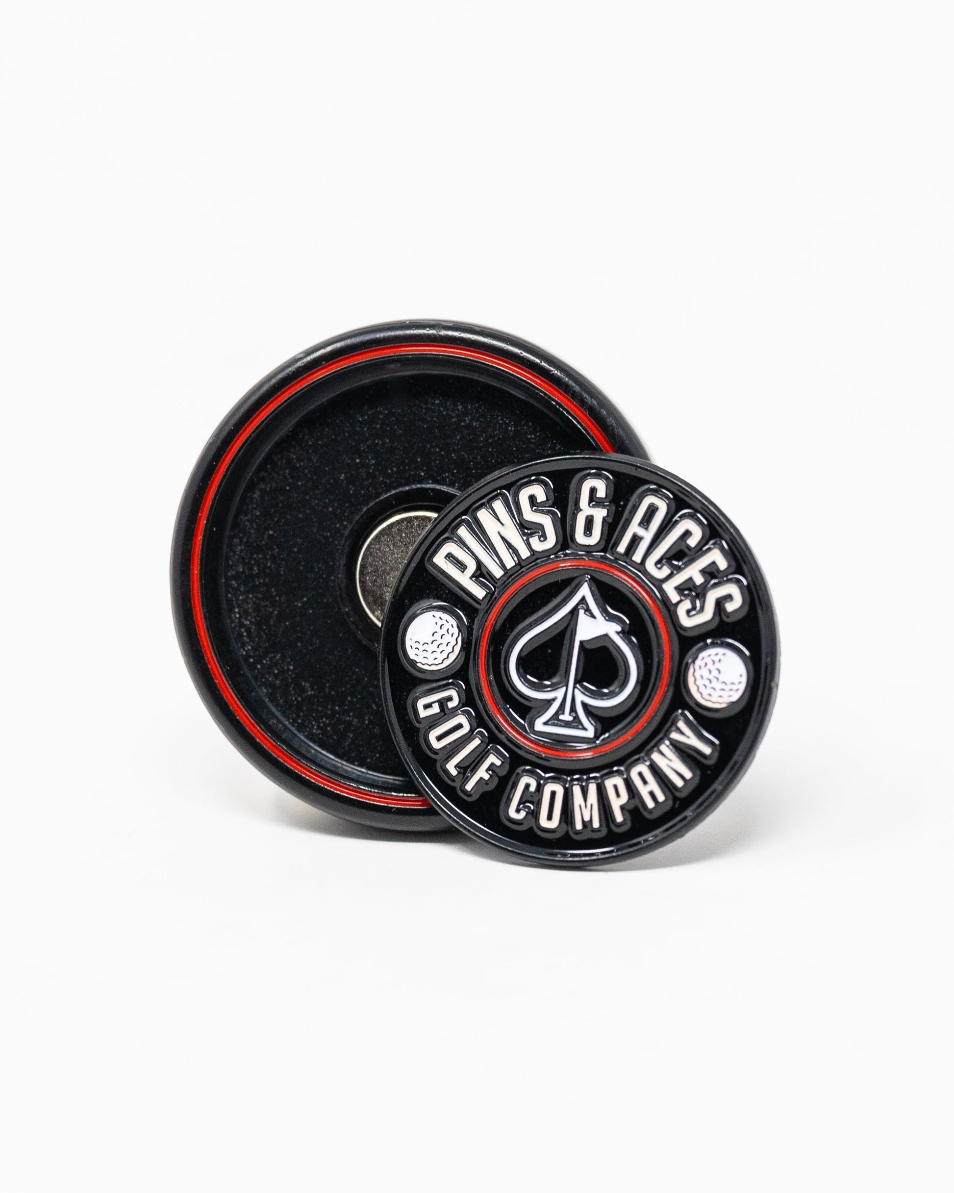 The Pop Out Medallion Ball Marker - Black by Pins and Aces features a round, black, magnetic marker with white branding, two golf balls, and an ace of spades symbol. It includes a thin marker displayed in front of its round holder with a red ring.