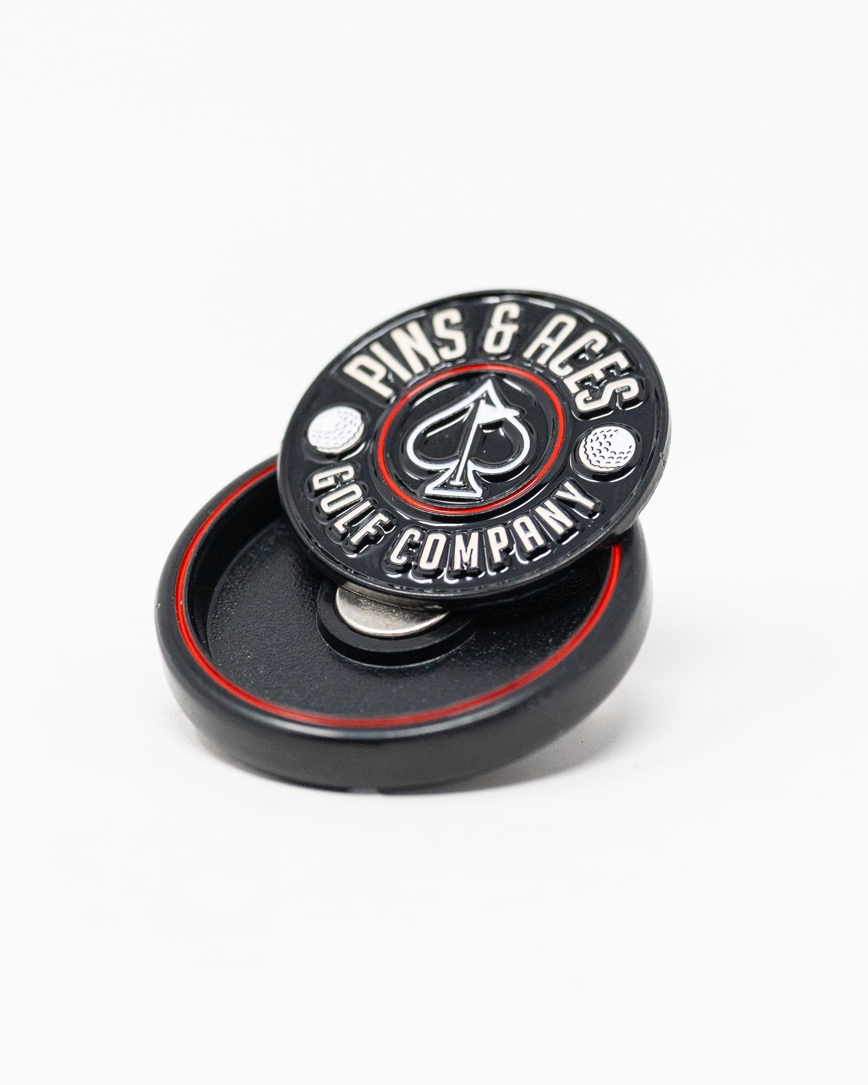 The Pop Out Medallion Ball Marker - Black by Pins and Aces, featuring an ace of spades logo, sits on its matching magnetic holder. This black and red golf accessory is displayed against a white background.