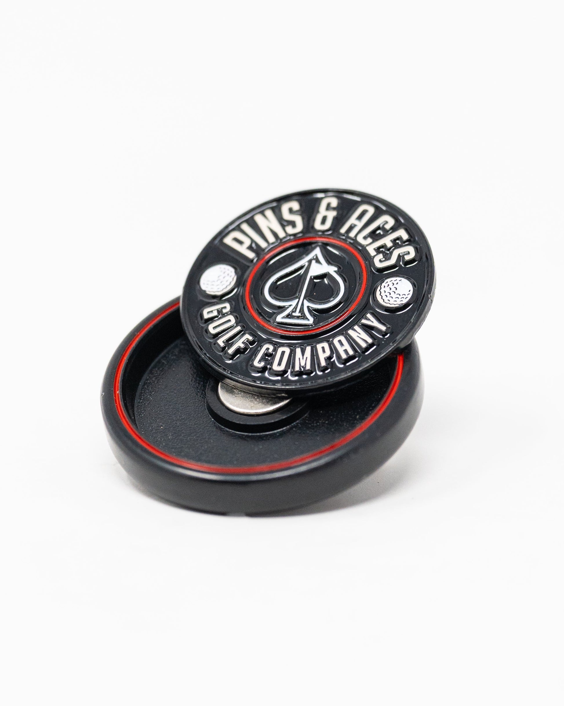 The Pop Out Medallion Ball Marker - Black by Pins and Aces, featuring an ace of spades logo, sits on its matching magnetic holder. This black and red golf accessory is displayed against a white background.