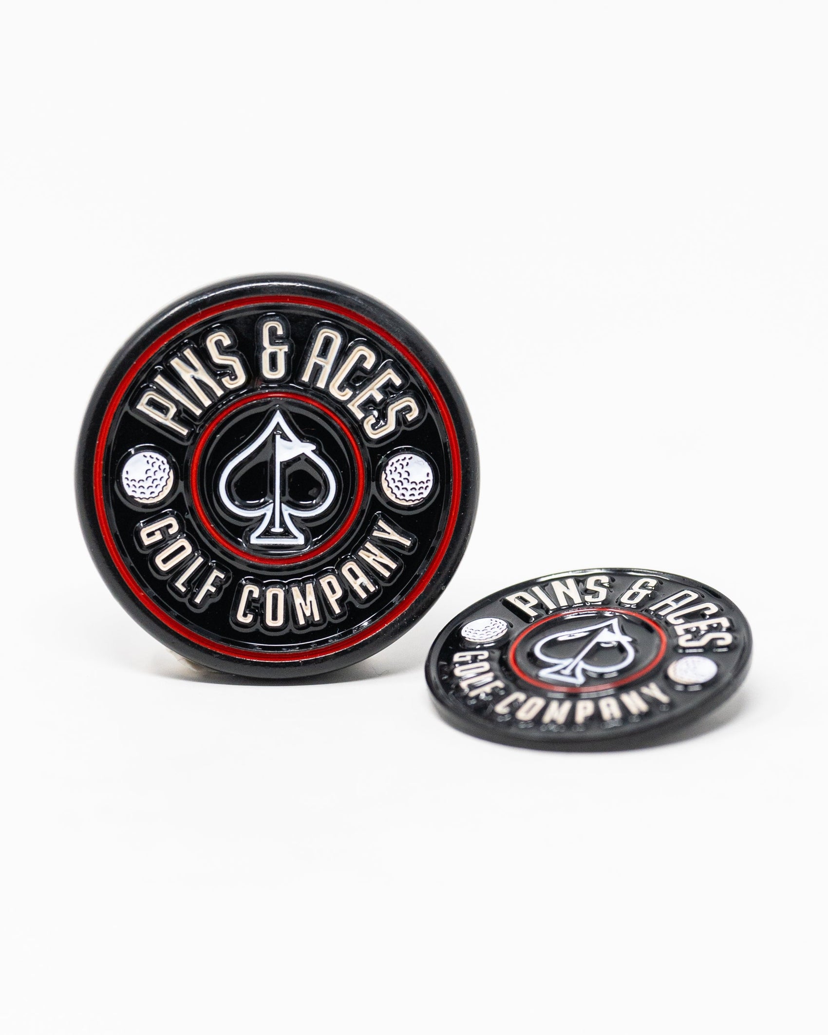 The Pop Out Medallion Ball Marker - Black by Pins and Aces features round black and red ball markers with ace of spades symbol; one stands upright while the other lies flat.