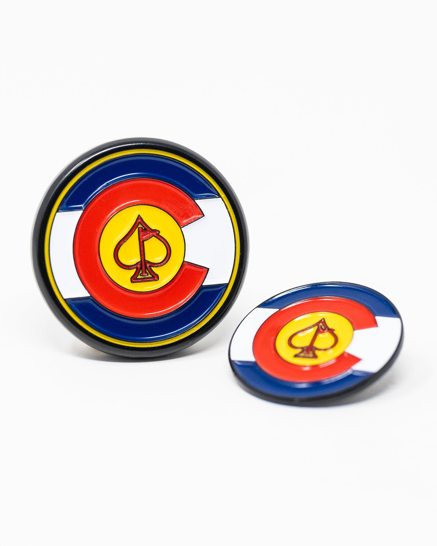 The Pins and Aces Pop Out Medallion Ball Marker - Colorado features two round markers with a Colorado flag design, including a yellow spade inside a red "C" on blue, white, and red backgrounds—one upright, one flat.