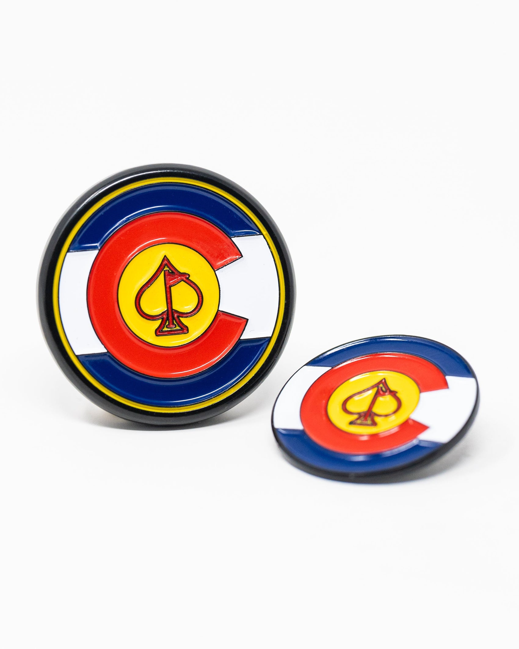 The Pins and Aces Pop Out Medallion Ball Marker - Colorado features two round markers with a Colorado flag design, including a yellow spade inside a red "C" on blue, white, and red backgrounds—one upright, one flat.
