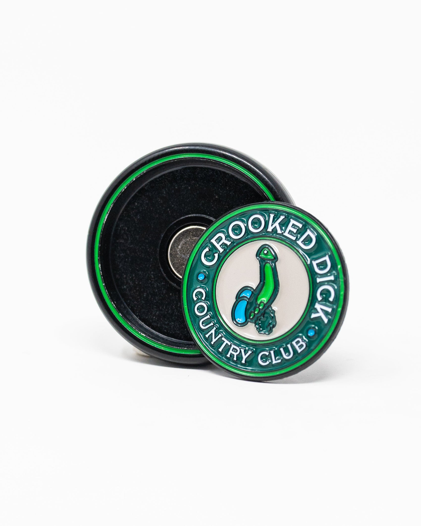 The Pins and Aces Pop Out Medallion Ball Marker - Crooked Dick displays "CROOKED DICK COUNTRY CLUB" around a cartoon green phallic golf club, shown positioned in front of an open black case.