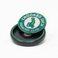 The Pop Out Medallion Ball Marker - Crooked Dick by Pins and Aces features "CROOKED DICK COUNTRY CLUB" encircling a cartoon bent green golf club with two blue balls, and comes in a sleek black round case.