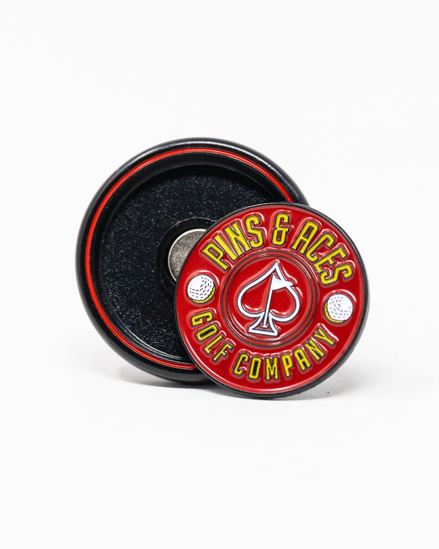 The Pop Out Medallion Ball Marker - Red by Pins and Aces features a round red marker with “Pins & Aces Golf Company” around an ace of spades and golf balls, displayed next to its black magnetic holder.