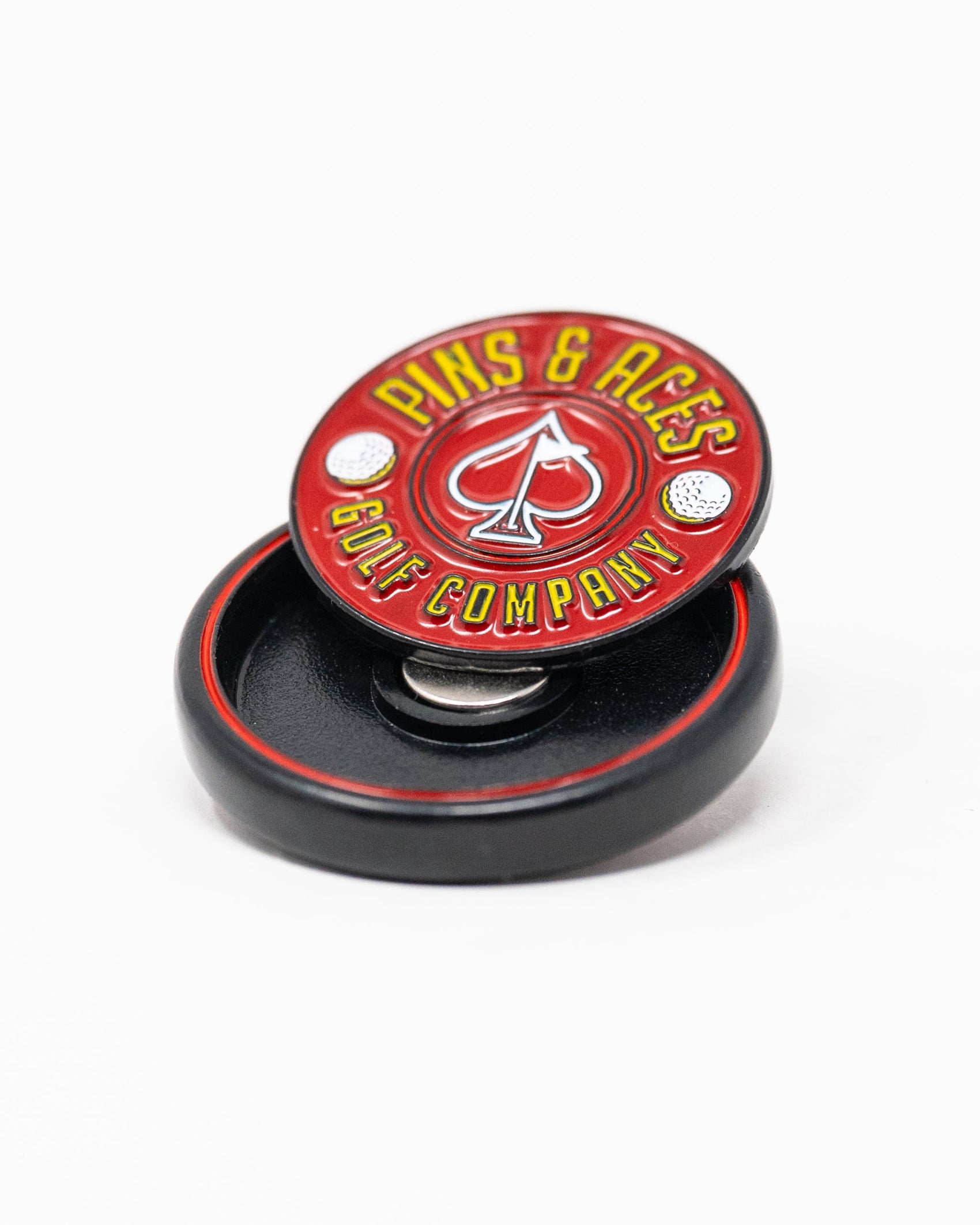 The Pop Out Medallion Ball Marker - Red by Pins and Aces features a round, red design with company text and an ace of spades symbol, shown upright in a black circular case against a white background.