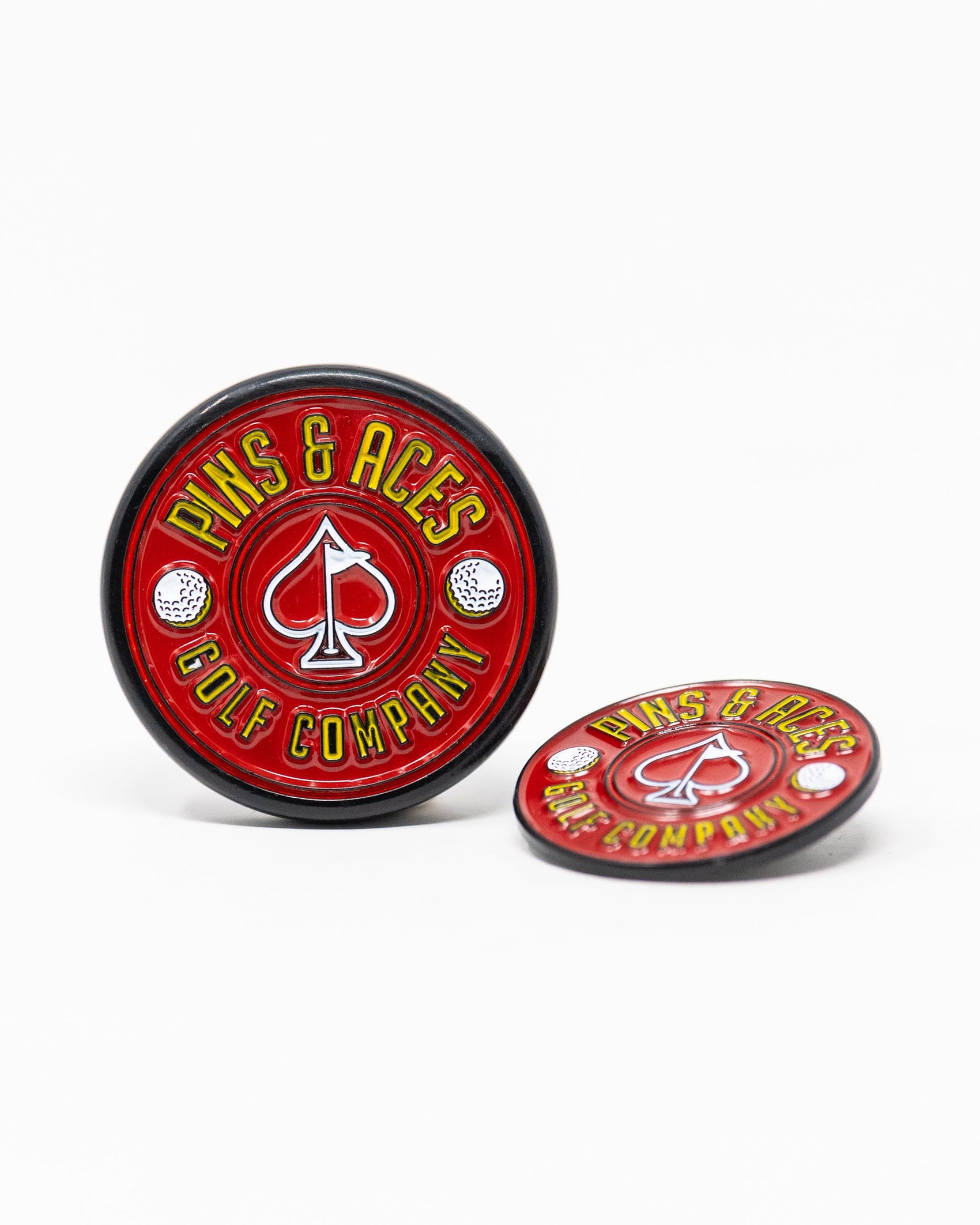 The Pop Out Medallion Ball Marker - Red by Pins and Aces features “PINS AND ACES” in yellow around a spade and two golf balls; one magnetic marker stands upright while the other lies flat.
