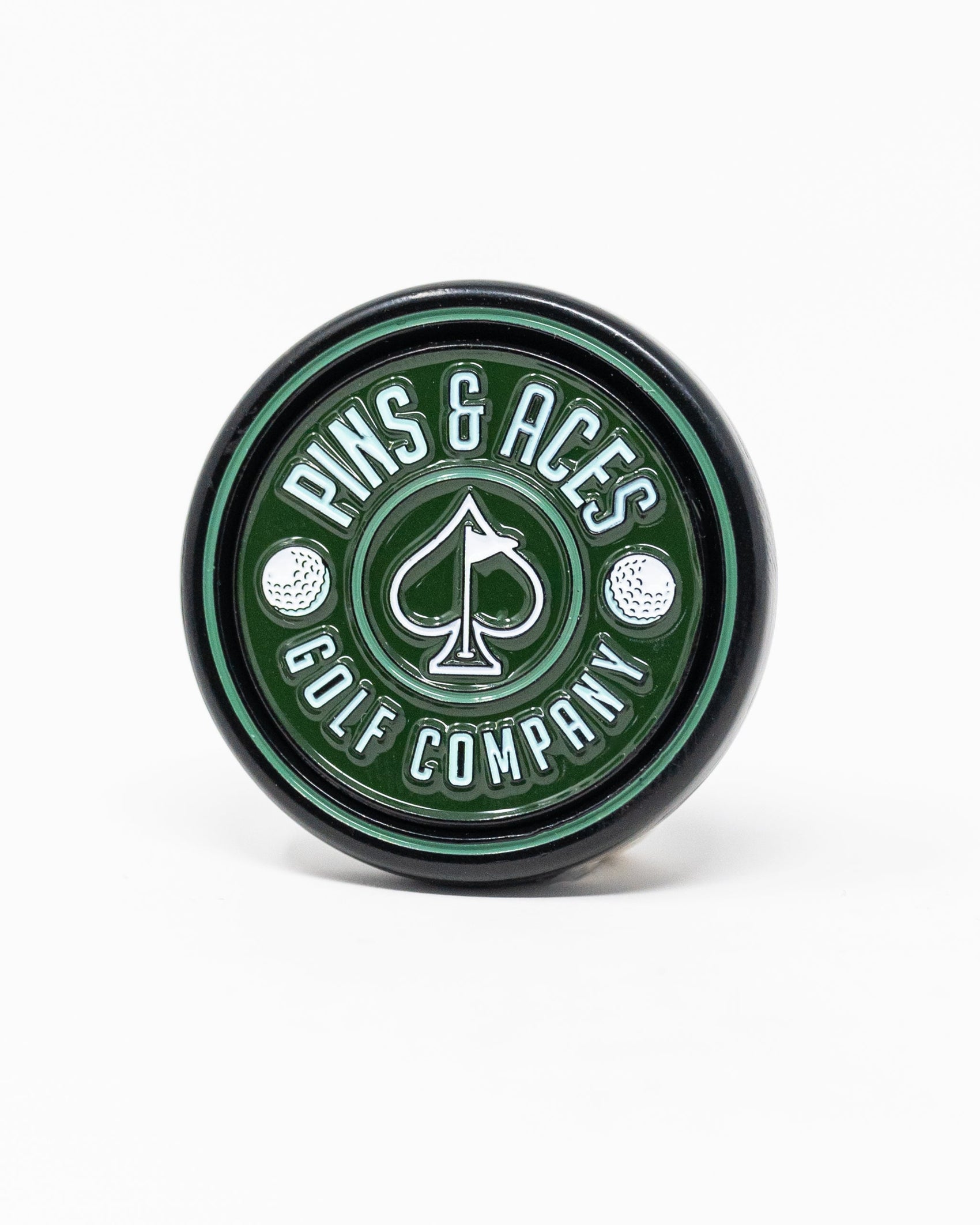 The Pop Out Medallion Ball Marker - Green by Pins and Aces is a round green and black golf accessory featuring the brand name around a spade symbol and two golf balls on a white background.