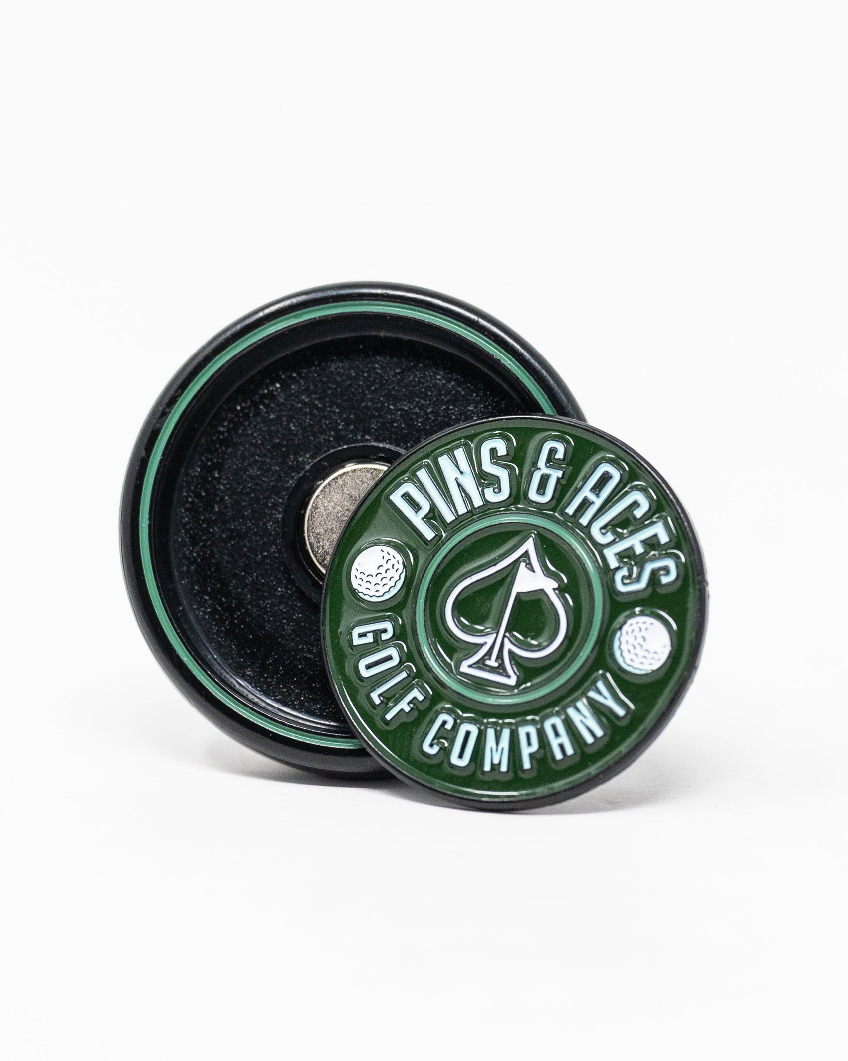 The Pop Out Medallion Ball Marker - Green by Pins and Aces is a round green and black magnetic golf accessory, featuring "Pins and Aces" with an illustrated spade, shown upright with its magnetic back displayed flat behind it.