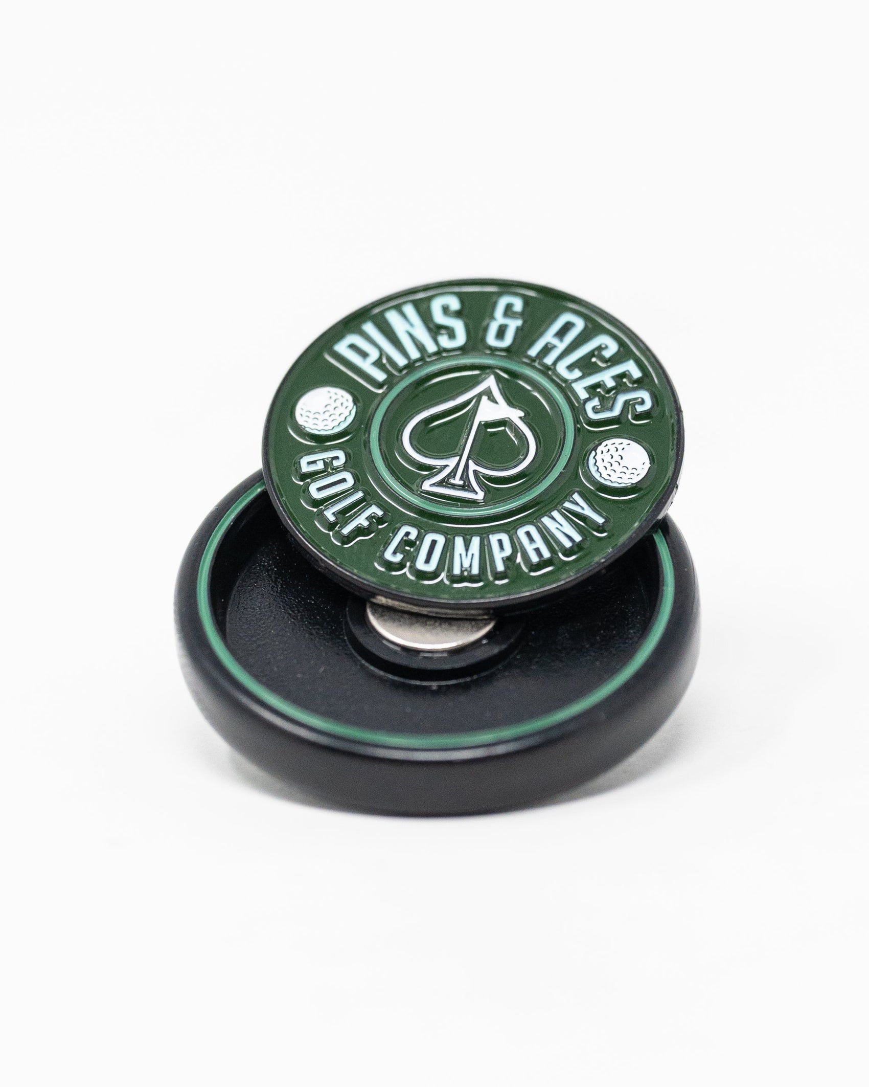 The Pop Out Medallion Ball Marker - Green by Pins and Aces, shown with two golf balls on a small black round holder, is an essential golf accessory displayed against a white background.