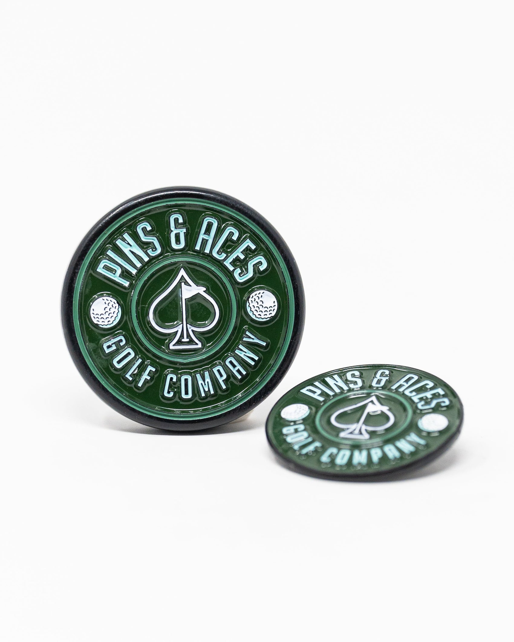 Two green Pop Out Medallion Ball Markers by Pins and Aces, each with a white spade symbol and branding; one stands upright while the other lies flat, showcasing a stylish golf accessory on a white background.