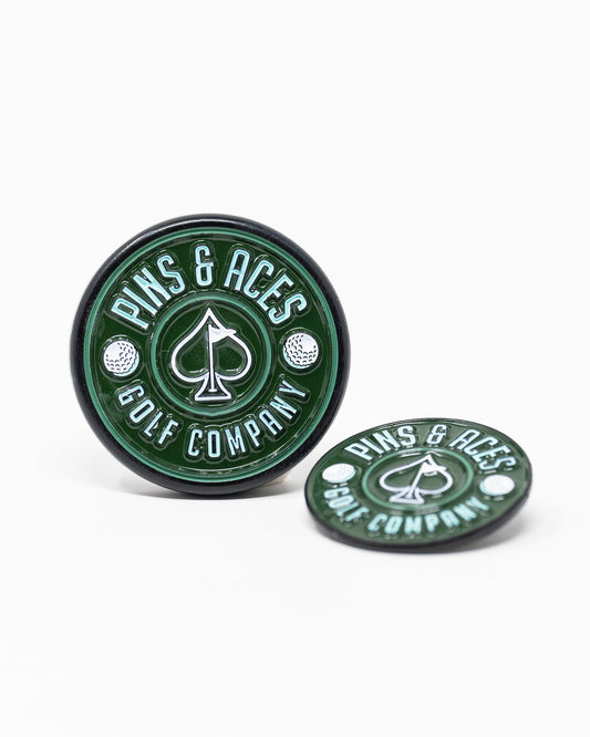Two green Pop Out Medallion Ball Markers by Pins and Aces, each with a white spade symbol and branding; one stands upright while the other lies flat, showcasing a stylish golf accessory on a white background.