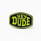 The Pins and Aces HEYDUDE - Logo Ball Marker is a black enamel pin that doubles as a cast metal ball marker, displaying HEY DUDE in bold yellow-green letters with white trim on a simple white background.
