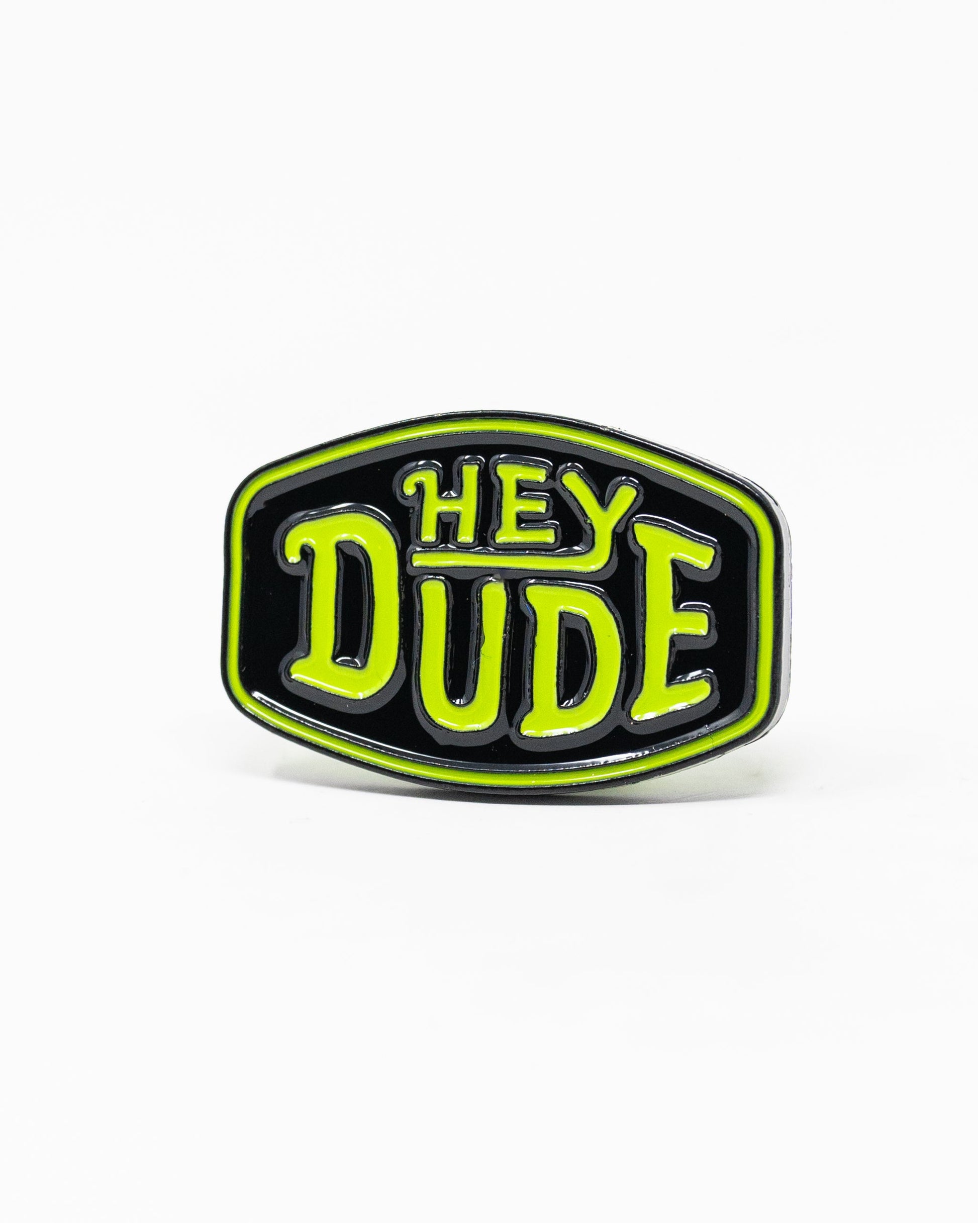 The Pins and Aces HEYDUDE - Logo Ball Marker is a black enamel pin that doubles as a cast metal ball marker, displaying HEY DUDE in bold yellow-green letters with white trim on a simple white background.
